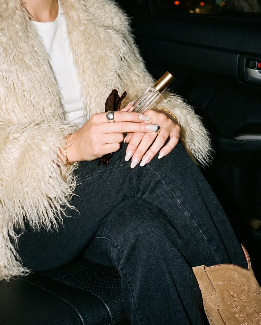 Hands with rings holding a Concrete Cowgirl perfume roller against a cream faux fur coat